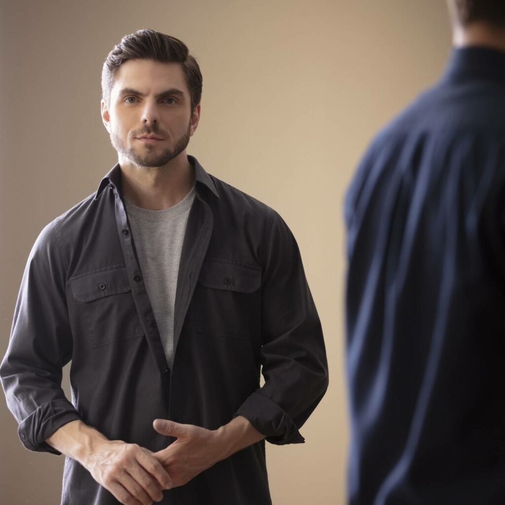 Homem em estúdio com fundo neutro vestindo camisa casual escura de manga longa sobre camiseta cinza, cabelos curtos e barba bem aparada, em enquadramento médio de frente com mãos unidas, transmitindo sobriedade, confiança e moda masculina contemporânea.