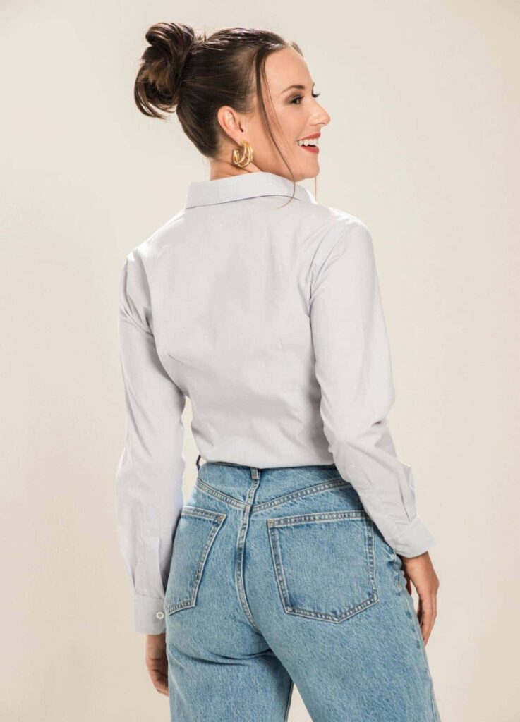 Mulher em estúdio com fundo claro vista de costas vestindo camisa social branca de manga longa e jeans azul de cintura alta, cabelo preso em coque e brincos dourados, em pose leve e elegante que destaca caimento da peça e estilo feminino contemporâneo.