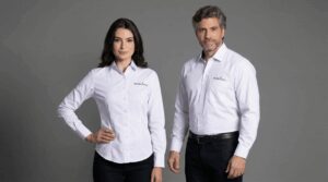 Dois profissionais vestindo camisas sociais corporativas brancas personalizadas com bordado da marca Decolar.com no lado esquerdo do peito. O modelo masculino usa camisa social branca de manga longa com bolso frontal e fechamento por botões, com o logo Decolar.com bordado em azul e vermelho acima do bolso. A modelo feminina veste camisa social branca de manga longa com modelagem acinturada, sem bolso, também com bordado Decolar.com no peito esquerdo. O fundo é preto, destacando as peças e a identidade visual da empresa do setor de turismo e viagens.