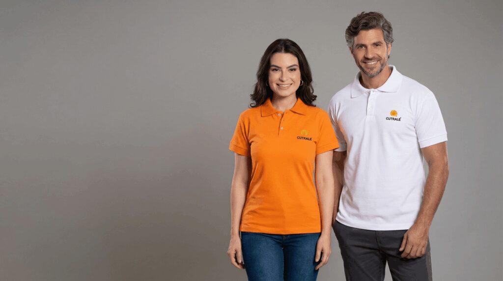 Dois profissionais vestindo polos corporativas personalizadas com bordado Cutrale no lado esquerdo do peito. A modelo feminina usa polo laranja com gola estruturada e peitilho com botões, apresentando o logo Cutrale em amarelo e azul. O modelo masculino veste polo branca com gola tradicional e bordado Cutrale no peito esquerdo. Ambas as peças possuem mangas curtas, modelagem confortável e acabamento alinhado. O fundo é preto, destacando as cores das polos e reforçando a identidade visual da empresa do setor agroindustrial.