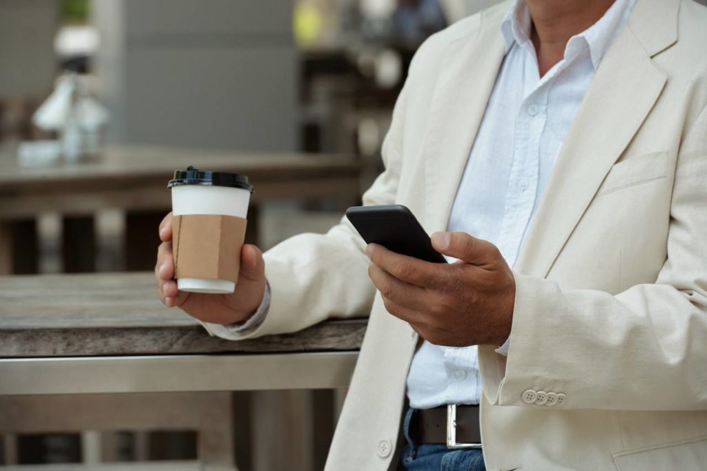 Homem de blazer claro tomando um cafe vendo o seu celular