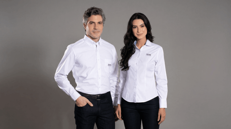 Dois profissionais, um homem e uma mulher, vestindo camisas sociais corporativas brancas da Sixtini. A camisa masculina possui bolso frontal no lado esquerdo com bordado discreto da marca “Sixtini Camisaria”, gola estruturada e fechamento por botões brancos. A camisa feminina apresenta modelagem acinturada, sem bolso, com bordado “Sixtini Camisaria” no lado esquerdo do peito e gola tradicional. Ambas são de manga longa, com caimento alinhado ao corpo. O fundo é azul escuro em degradê, destacando a elegância e a padronização do uniforme social corporativo.