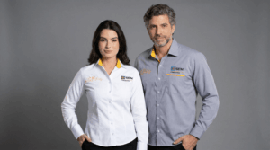 Dois profissionais, um homem e uma mulher, vestindo camisas sociais corporativas personalizadas com bordado da marca SEM. A modelo feminina usa camisa branca com detalhe interno amarelo na gola e bordado do logo SEM no lado esquerdo do peito, além de um bordado adicional em amarelo no lado direito. O modelo masculino veste camisa social cinza com detalhe amarelo interno na gola e bordado do logo SEM no peito esquerdo. Ambas as peças apresentam modelagem moderna, mangas longas e fechamento por botões frontais. O fundo é totalmente preto, destacando os uniformes e a identidade visual das camisas.