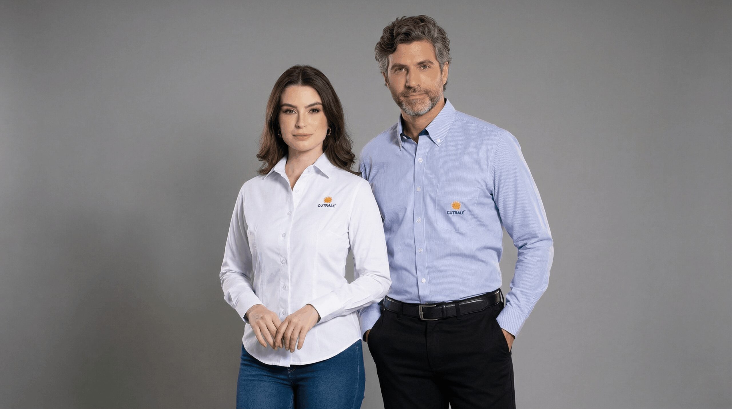 Dois profissionais, um homem e uma mulher, vestindo camisas sociais corporativas personalizadas com bordado da marca Cutrale no lado esquerdo do peito. A modelo feminina usa camisa social branca de manga longa, modelagem acinturada e fechamento frontal por botões. O modelo masculino veste camisa social azul clara de manga longa com bolso frontal e bordado Cutrale aplicado acima do bolso. O logo apresenta símbolo em laranja acompanhado do nome “Cutrale” em azul. O fundo é preto, destacando os uniformes e reforçando a identidade visual corporativa.