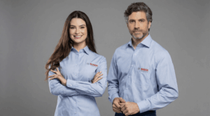 Dois profissionais, um homem e uma mulher, vestindo camisas sociais corporativas azul clara com padronagem discreta em listras finas. Ambas as camisas possuem bordado do logo BOSCH no lado esquerdo do peito, em vermelho com símbolo circular acima do nome. A modelo feminina está com os braços cruzados e a camisa acinturada, enquanto o modelo masculino veste camisa social masculina de corte tradicional com bolso frontal. O fundo é escuro e neutro, destacando os uniformes. Na parte inferior da imagem aparece a marca Sixtini est. 1992 e a frase “Artigo blog”.