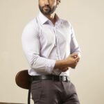 Homem em estúdio usando camisa social masculina em tom claro levemente lilás, mangas longas ajustadas, tecido liso e bem estruturado, combinada com calça escura e cinto preto, apoiado em um banco alto, fundo neutro bege, postura elegante que destaca o caimento e a sofisticação da peça.