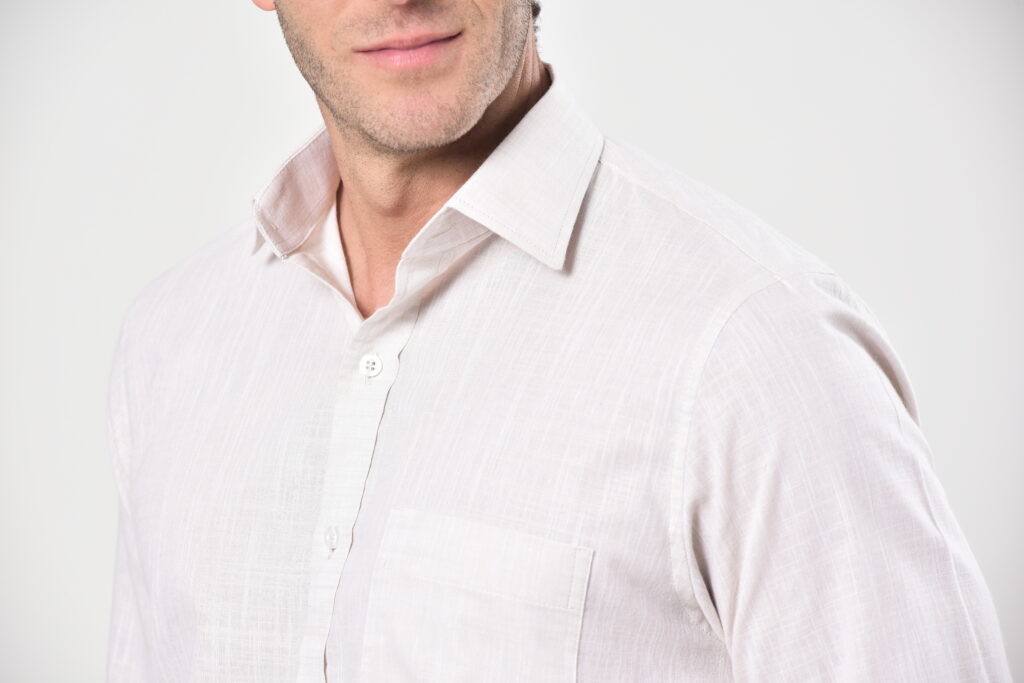 Close-up em estúdio de camisa social branca masculina em tecido leve e translúcido, destacando colarinho, botões e bolso frontal, com fundo claro, enfatizando textura, acabamento e elegância da moda masculina contemporânea.