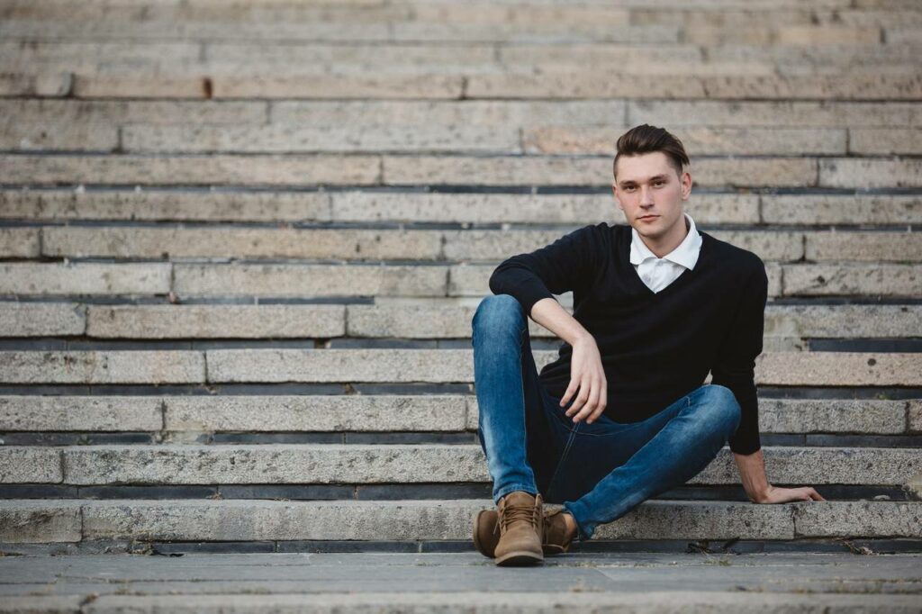 Homem sentado na escada de suéter e camisa branco por baixo de calça jeans