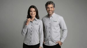 Dois profissionais vestindo camisas sociais corporativas cinza claras personalizadas com bordado Sicoob no lado esquerdo do peito. A camisa masculina possui bolso frontal e fechamento por botões pretos contrastantes, com o logo Sicoob em verde e azul aplicado acima do bolso. A camisa feminina apresenta modelagem acinturada, sem bolso, também com botões escuros e bordado Sicoob no peito esquerdo. Ambas são de manga longa e possuem acabamento refinado. O fundo é branco, destacando o uniforme e reforçando a identidade visual da cooperativa financeira.