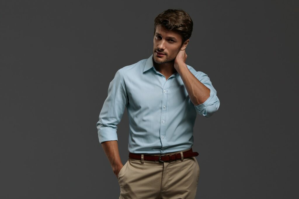 Homem em estúdio com fundo cinza escuro vestindo camisa social azul clara de manga longa e calça bege com cinto marrom, em enquadramento médio com uma mão no bolso e outra apoiada no pescoço, transmitindo sofisticação, estilo casual elegante e moda masculina contemporânea.