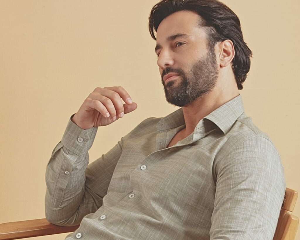 Homem adulto em ambiente interno com fundo bege vestindo camisa social bege de manga longa com textura leve, barba bem cuidada e cabelo escuro penteado para trás, em pose sentada e olhar contemplativo que transmite sofisticação, elegância casual e moda masculina contemporânea.