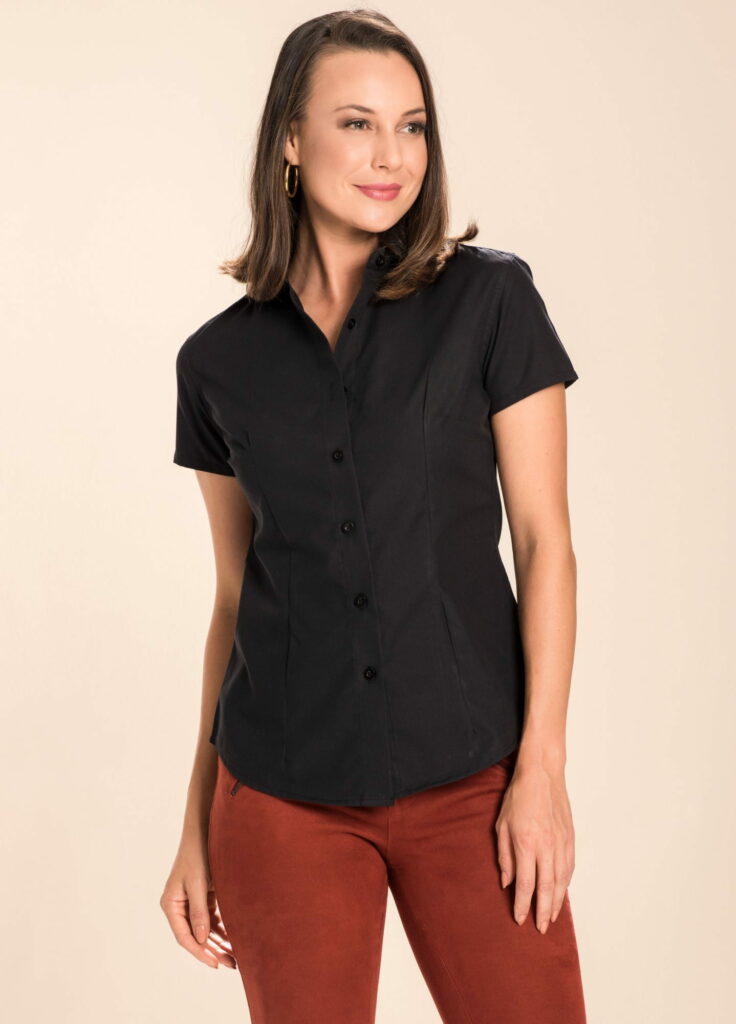 Mulher em estúdio com fundo claro vestindo camisa social preta de manga curta e calça marrom avermelhada, cabelo solto na altura dos ombros e brincos dourados, em pose elegante com olhar lateral que transmite sofisticação, versatilidade e moda feminina profissional contemporânea.