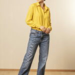 Mulher em estúdio com fundo claro vestindo camisa social amarela de manga longa e jeans azul de corte reto, usando óculos escuros, cabelo preso e tênis casuais, em pose de corpo inteiro descontraída que transmite estilo urbano, elegância casual e moda feminina contemporânea.