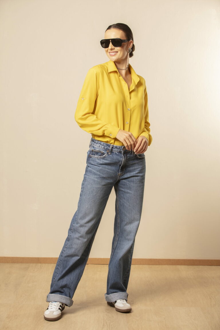 Mulher em estúdio com fundo claro vestindo camisa social amarela de manga longa e jeans azul de corte reto, usando óculos escuros, cabelo preso e tênis casuais, em pose de corpo inteiro descontraída que transmite estilo urbano, elegância casual e moda feminina contemporânea.