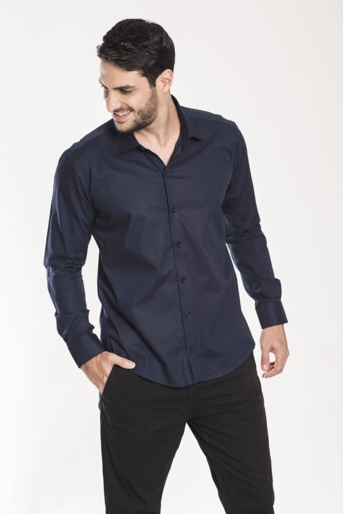 Homem jovem em estúdio com fundo claro vestindo camisa social azul marinho de manga longa e calça preta, em pose descontraída com a mão no bolso e olhar lateral, transmitindo elegância casual, moda masculina contemporânea e estilo profissional versátil.