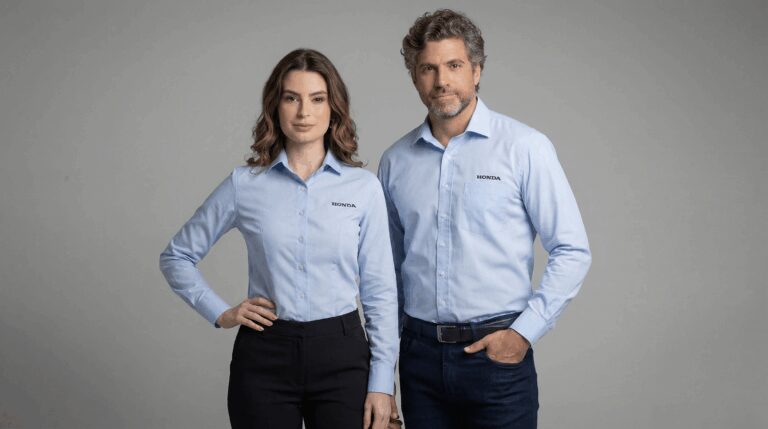 Dois profissionais vestindo camisas sociais corporativas azul clara personalizadas com bordado Honda no lado esquerdo do peito. O modelo masculino usa camisa de manga longa com bolso frontal e bordado “HONDA” aplicado acima do bolso. A modelo feminina veste camisa social azul clara de manga longa com modelagem acinturada e bordado Honda no peito esquerdo, sem bolso. Ambas as peças possuem fechamento frontal por botões e acabamento alinhado. O fundo é branco, destacando a tonalidade clara das camisas e reforçando a identidade visual da marca automotiva.