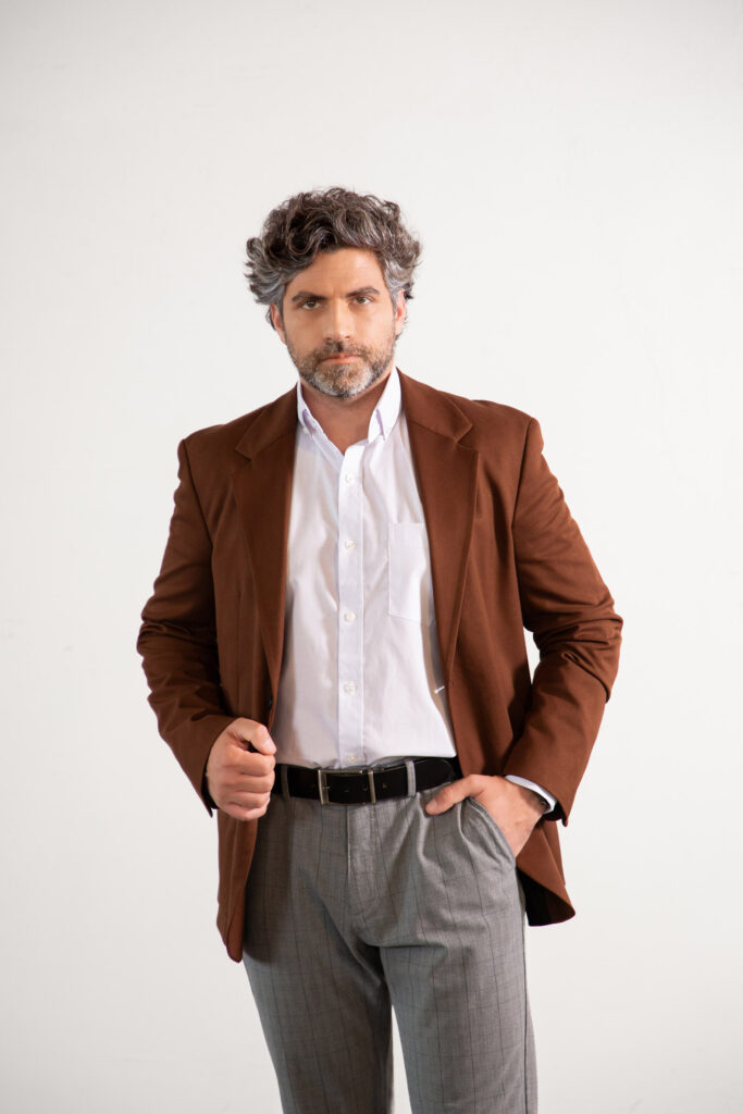 Homem adulto com cabelo grisalho e barba curta vestindo blazer marrom, camisa social branca e calça cinza, em pose de corpo quase inteiro com uma mão no bolso, fotografado em estúdio com fundo claro, transmitindo sofisticação, estilo executivo e moda masculina contemporânea.