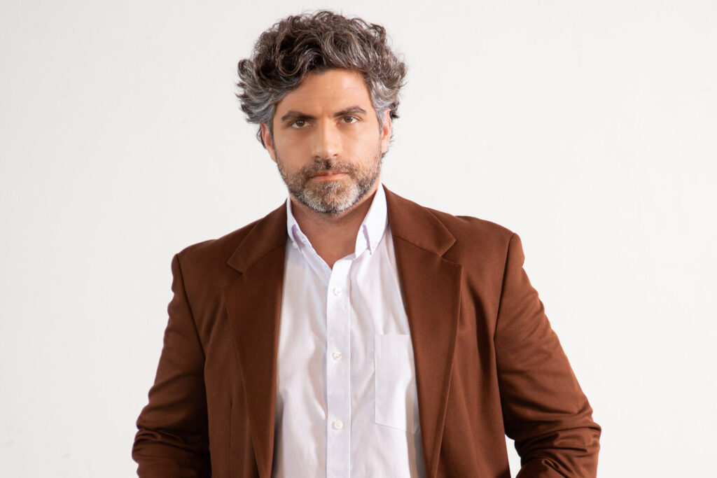 Homem adulto com cabelo grisalho e barba curta vestindo blazer marrom e camisa social branca, em retrato de estúdio com fundo claro e iluminação suave, transmitindo elegância, maturidade e estilo corporativo contemporâneo.