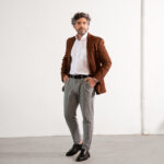 Homem adulto em estúdio com fundo claro vestindo blazer marrom, camisa social branca e calça cinza, em pose de corpo inteiro com as mãos nos bolsos, composição minimalista que transmite elegância, sofisticação e moda masculina profissional contemporânea.