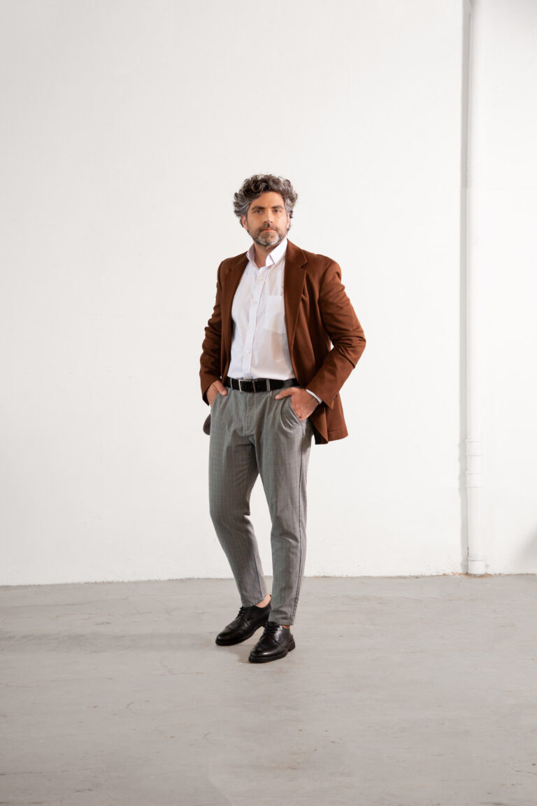 Homem adulto em estúdio com fundo claro vestindo blazer marrom, camisa social branca e calça cinza, em pose de corpo inteiro com as mãos nos bolsos, composição minimalista que transmite elegância, sofisticação e moda masculina profissional contemporânea.