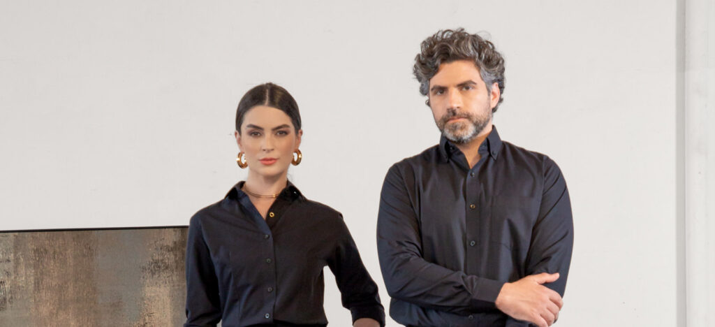 Homem e mulher lado a lado em estúdio com fundo claro vestindo camisas sociais pretas, ela com acessórios dourados e ele com braços cruzados, enquadramento médio que transmite sobriedade, elegância, moda corporativa contemporânea e posicionamento profissional premium.