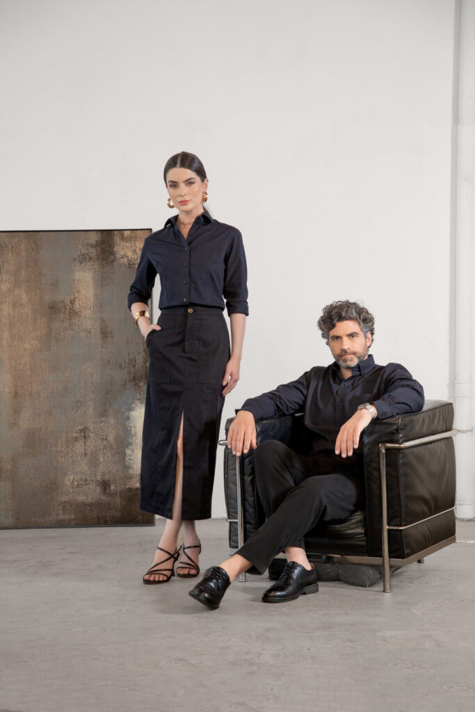 Homem e mulher em estúdio com fundo neutro vestindo looks monocromáticos em preto, ela em pé com camisa social e saia midi com fenda e ele sentado em poltrona de couro usando camisa social e calça preta, composição sofisticada que transmite elegância, moda contemporânea e estilo profissional premium.