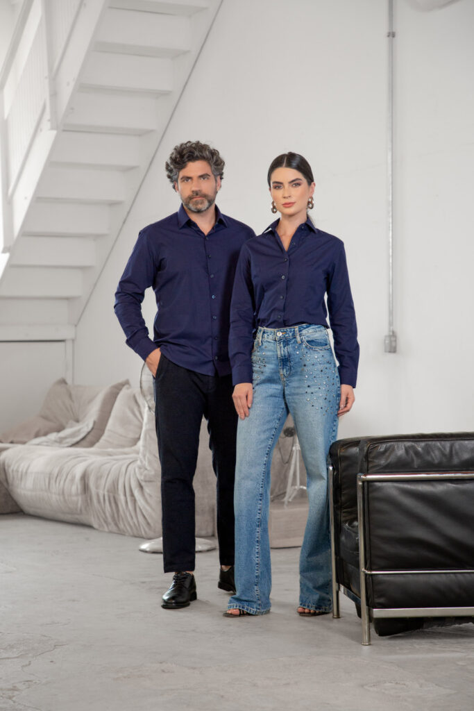 Homem e mulher em ambiente interno moderno vestindo camisas sociais azul marinho de manga longa, ele com calça preta e sapato social e ela com jeans azul de cintura alta e aplicação de pedrarias, em cenário clean com sofá e escada ao fundo, destacando elegância casual e moda contemporânea.