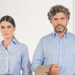 Homem e mulher em ambiente interno claro vestindo camisas sociais azul claro, ela com acessórios discretos e ele segurando um blazer bege, enquadramento médio com escada ao fundo que transmite profissionalismo, elegância corporativa e estilo contemporâneo para ambientes de trabalho.