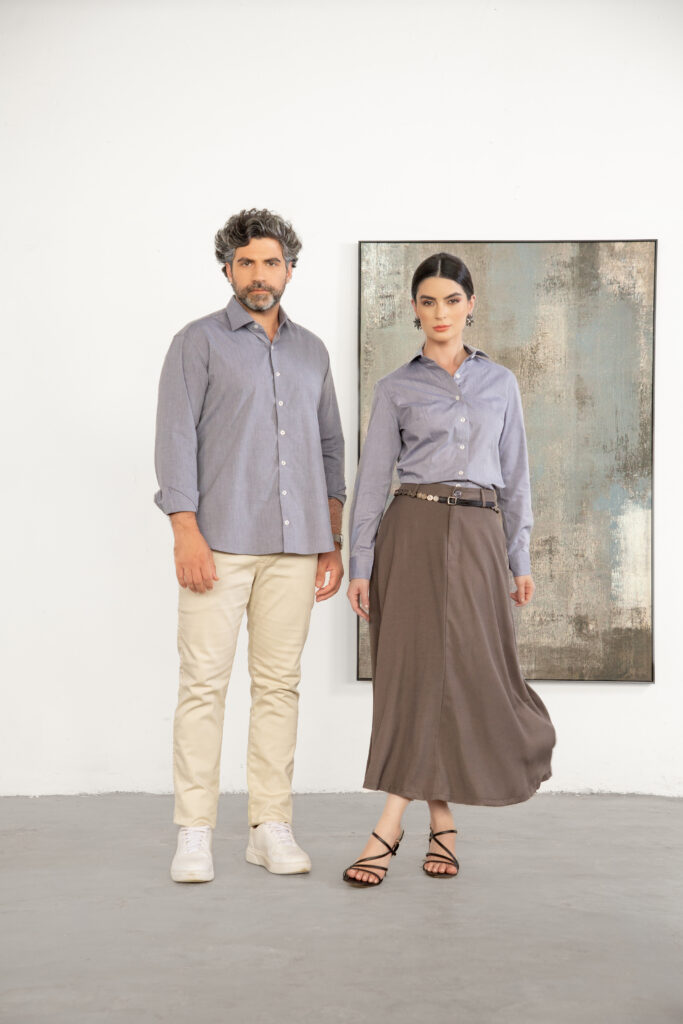 Homem e mulher em estúdio com fundo claro vestindo camisas sociais cinza, ele com calça bege e tênis claros e ela com saia midi marrom e sandálias, composição clean e equilibrada que transmite elegância casual, moda contemporânea e versatilidade para ambiente profissional.