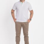 Imagem em estúdio com fundo branco mostrando um homem adulto em pose frontal, de corpo inteiro. Ele veste uma camisa polo branca de manga curta, com gola estruturada e detalhe interno em tom verde claro na parte interna da carcela. A polo tem caimento reto e aparência limpa, típica de fotografia de catálogo. O modelo usa calça chino em tom bege ou marrom claro, com as mãos apoiadas nos bolsos frontais, e sapatos pretos de couro com acabamento clássico. No pulso direito, usa um relógio metálico. Possui barba curta bem aparada, cabelos ondulados com fios grisalhos e expressão amigável com leve sorriso. Há tatuagens visíveis nos antebraços. A iluminação é uniforme e suave, destacando a roupa e mantendo um visual neutro e profissional.