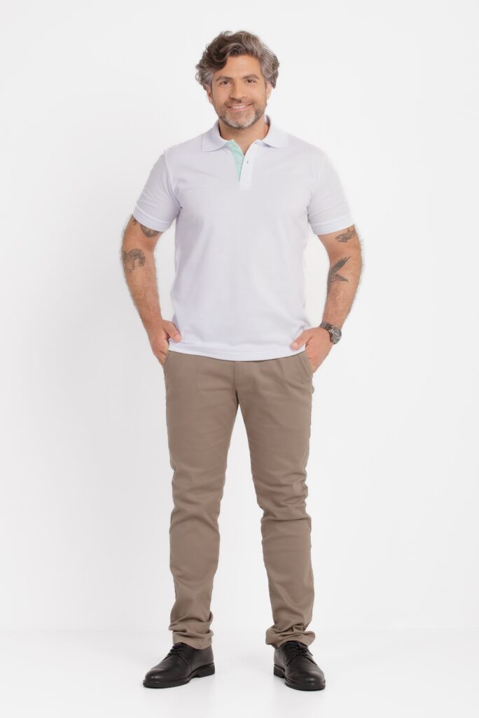 Imagem em estúdio com fundo branco mostrando um homem adulto em pose frontal, de corpo inteiro. Ele veste uma camisa polo branca de manga curta, com gola estruturada e detalhe interno em tom verde claro na parte interna da carcela. A polo tem caimento reto e aparência limpa, típica de fotografia de catálogo. O modelo usa calça chino em tom bege ou marrom claro, com as mãos apoiadas nos bolsos frontais, e sapatos pretos de couro com acabamento clássico. No pulso direito, usa um relógio metálico. Possui barba curta bem aparada, cabelos ondulados com fios grisalhos e expressão amigável com leve sorriso. Há tatuagens visíveis nos antebraços. A iluminação é uniforme e suave, destacando a roupa e mantendo um visual neutro e profissional.