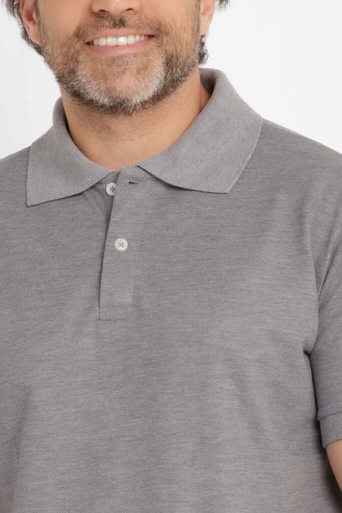 Imagem em estúdio com fundo branco mostrando um close do tronco e parte inferior do rosto de um homem adulto. Ele veste uma camisa polo cinza claro mesclado, com gola tradicional estruturada e carcela frontal com dois botões claros. O tecido apresenta textura fina e uniforme, típica de malha piquê, com aspecto suave e caimento confortável. A barba é curta e bem aparada, com fios grisalhos misturados ao castanho, e há um leve sorriso visível. A iluminação é difusa e equilibrada, destacando a cor e a textura da camisa sem sombras marcadas, reforçando um visual limpo e próprio para fotografia de catálogo de moda masculina.