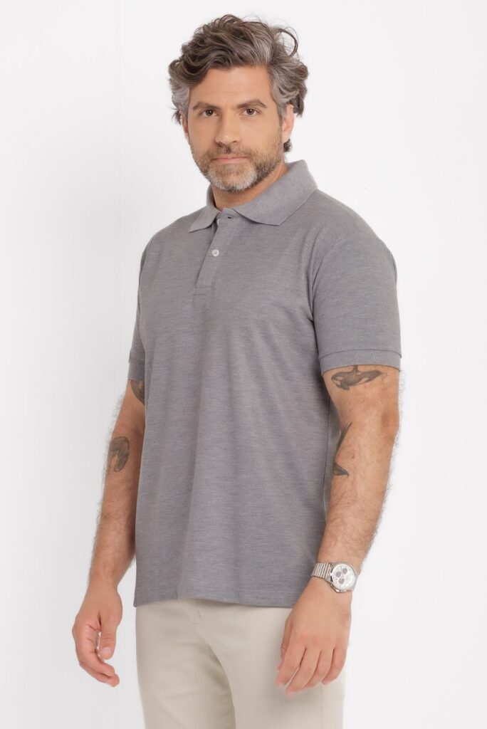 Imagem em estúdio com fundo branco e iluminação uniforme, mostrando um homem adulto em plano médio, levemente de perfil. Ele veste uma camisa polo cinza claro mesclado, de manga curta, com gola tradicional e carcela discreta com botões claros. O tecido tem aparência lisa e o caimento é reto, confortável, sem ajuste excessivo ao corpo. O modelo usa calça clara em tom bege ou off white e um relógio metálico prateado no pulso esquerdo. Os braços estão relaxados ao lado do corpo, com tatuagens visíveis nos antebraços. O cabelo é ondulado, castanho com fios grisalhos, volumoso e bem arrumado. A barba é curta e bem aparada. A expressão facial é neutra e confiante, com olhar direcionado para a câmera. A composição transmite um estilo clean, casual e elegante, típico de fotografia de catálogo de moda masculina.