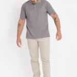 Imagem em plano quase inteiro de um homem adulto em estúdio, com fundo totalmente branco e iluminação uniforme. Ele veste uma camisa polo de cor cinza claro mesclado, com gola tradicional e carcela frontal discreta. O caimento é reto, confortável, acompanhando o corpo sem ficar ajustado. O modelo está usando calça clara em tom bege ou off white e tênis branco de visual minimalista. Ele olha para baixo, com a cabeça levemente inclinada, criando uma postura casual e natural. Os braços estão relaxados ao lado do corpo, revelando tatuagens nos antebraços e um relógio metálico no pulso esquerdo. O cabelo é ondulado, em tom castanho com fios grisalhos, e a barba é curta e bem aparada. A composição geral transmite um estilo clean, casual e elegante, típico de fotografia de catálogo de moda.