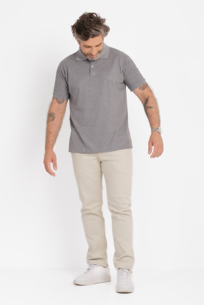 Imagem em plano quase inteiro de um homem adulto em estúdio, com fundo totalmente branco e iluminação uniforme. Ele veste uma camisa polo de cor cinza claro mesclado, com gola tradicional e carcela frontal discreta. O caimento é reto, confortável, acompanhando o corpo sem ficar ajustado. O modelo está usando calça clara em tom bege ou off white e tênis branco de visual minimalista. Ele olha para baixo, com a cabeça levemente inclinada, criando uma postura casual e natural. Os braços estão relaxados ao lado do corpo, revelando tatuagens nos antebraços e um relógio metálico no pulso esquerdo. O cabelo é ondulado, em tom castanho com fios grisalhos, e a barba é curta e bem aparada. A composição geral transmite um estilo clean, casual e elegante, típico de fotografia de catálogo de moda.