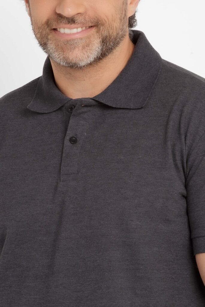 Imagem em close do tronco e parte inferior do rosto de um homem adulto em estúdio. Ele veste uma camisa polo de cor cinza escuro mesclado, com gola tradicional e carcela frontal com dois botões pretos visíveis. O tecido apresenta textura típica de malha polo, com aspecto levemente granulado e uniforme. É possível ver parte do sorriso do modelo e a barba curta, com fios escuros e grisalhos. O caimento da camisa é reto, acompanhando o corpo sem marcar excessivamente. O fundo é totalmente branco, com iluminação homogênea, destacando a cor e a textura do tecido.