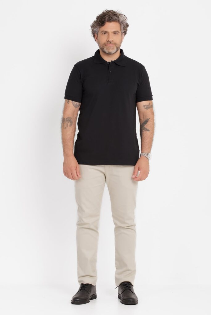 Imagem de estúdio em fundo branco mostrando um homem adulto em enquadramento de corpo inteiro, posicionado de frente para a câmera. Ele veste uma camisa polo preta lisa, de malha piquê, com gola estruturada e carcela curta com botões discretos no mesmo tom. A peça tem caimento regular, mangas curtas ajustadas e comprimento alinhado ao quadril, transmitindo um visual clássico e versátil. O modelo usa calça clara, em tom bege ou off-white, de corte reto e ajuste confortável, combinada com sapatos sociais pretos de couro. Ele possui cabelo ondulado com fios grisalhos, barba curta também grisalha e expressão neutra e confiante. Tatuagens são visíveis nos dois braços e ele usa um relógio metálico no pulso direito. A iluminação é uniforme e suave, típica de fotografia de catálogo, destacando o caimento da roupa e a textura do tecido sem sombras marcadas.