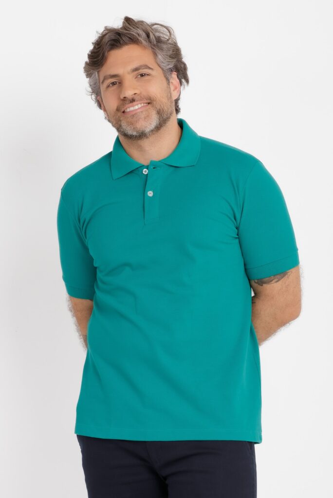 Imagem de estúdio em fundo branco mostrando um homem adulto em enquadramento médio, do topo da cabeça até o quadril. Ele veste uma camisa polo em tom verde água, de malha piquê, com gola estruturada no mesmo tom e carcela frontal com dois botões claros. A peça tem caimento regular, mangas curtas ajustadas e barra reta. O modelo está levemente inclinado para a direita, com as mãos posicionadas atrás do corpo, transmitindo postura relaxada. Possui cabelo ondulado com fios grisalhos, barba curta também grisalha e expressão amigável, com leve sorriso voltado para a câmera. Parte de tatuagens é visível nos braços. A iluminação é uniforme e suave, típica de fotografia de catálogo, destacando cor, textura e acabamento da camisa.