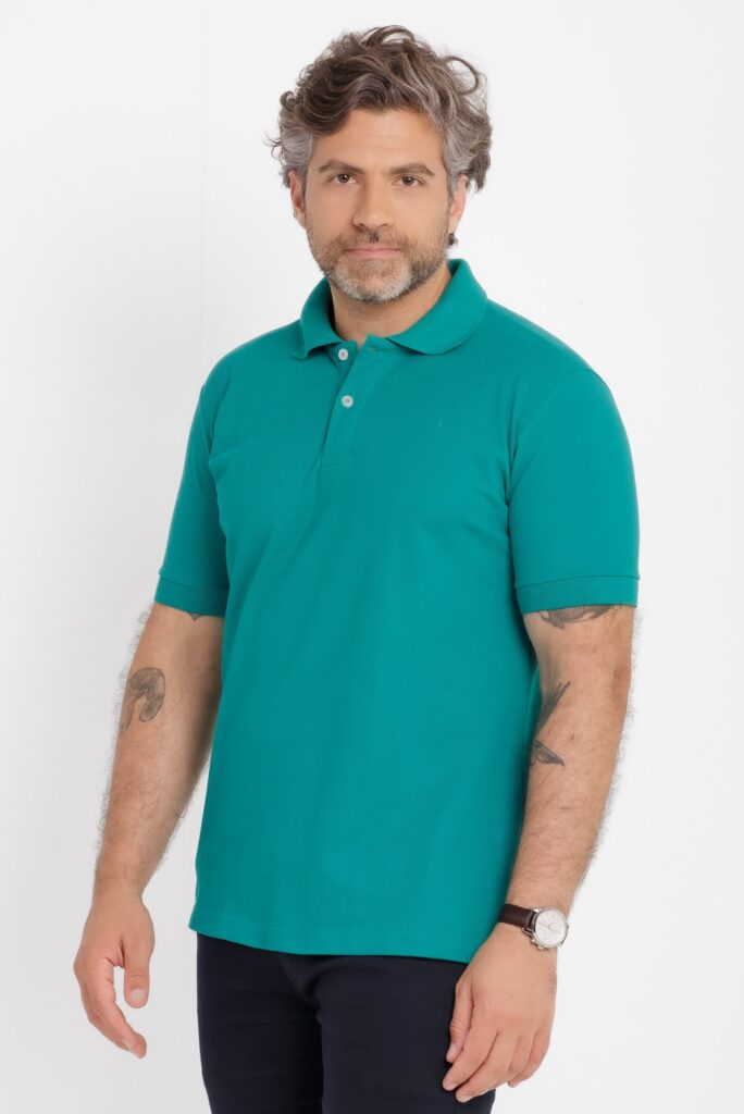 Imagem de estúdio em fundo branco mostrando um homem adulto em enquadramento médio, do topo da cabeça até parte das coxas. Ele veste uma camisa polo em tom verde água, confeccionada em malha piquê, com gola estruturada no mesmo tom e carcela frontal com dois botões claros. O caimento é regular, com mangas curtas ajustadas e barra reta. O modelo tem cabelo ondulado com fios grisalhos, barba curta também grisalha e expressão neutra, olhando diretamente para a câmera. Nos braços, há tatuagens visíveis e, no pulso direito, um relógio de caixa clara com pulseira escura. Iluminação uniforme e suave, típica de fotografia de catálogo de vestuário, destacando cor, textura e acabamento da peça.