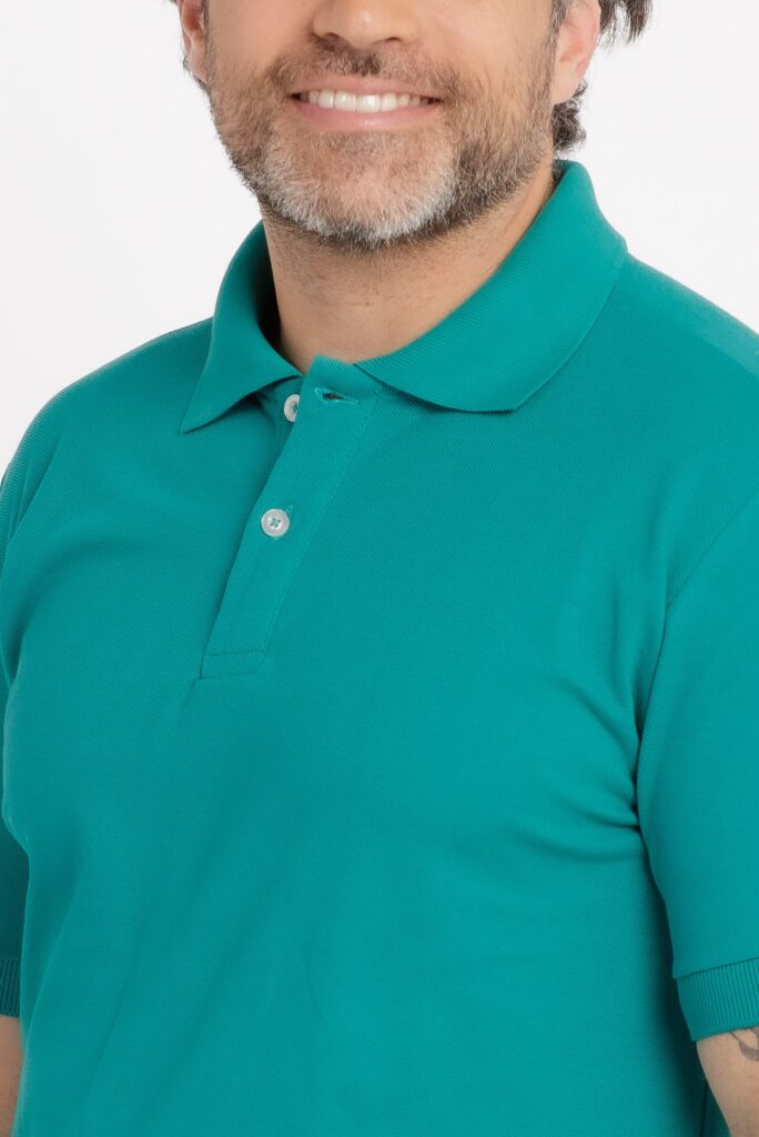 Close do tronco e parte inferior do rosto de um homem adulto em estúdio, com fundo branco. A imagem destaca uma camisa polo em tom verde água intenso, confeccionada em malha piquê com textura bem definida. A gola é estruturada, no mesmo tom da peça, e a carcela frontal possui dois botões brancos visíveis. O caimento é ajustado ao corpo, com a costura do ombro e da manga aparecendo com nitidez. O modelo apresenta barba curta com fios grisalhos e um leve sorriso, visível apenas na região da boca. Iluminação uniforme e limpa, típica de fotografia de catálogo de vestuário, com foco em acabamento, cor e textura do tecido.