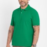 Homem adulto fotografado em estúdio com fundo branco, em enquadramento de meio corpo. Ele veste camisa polo verde de manga curta, com gola tradicional e carcela com dois botões claros, confeccionada em malha piquê de textura visível e caimento reto. O modelo tem cabelo ondulado com fios grisalhos, barba curta grisalha e expressão leve e confiante. Usa calça em tom bege e relógio metálico no pulso. Tatuagens discretas aparecem nos antebraços. Iluminação uniforme e estética clean de fotografia de catálogo de moda masculina.