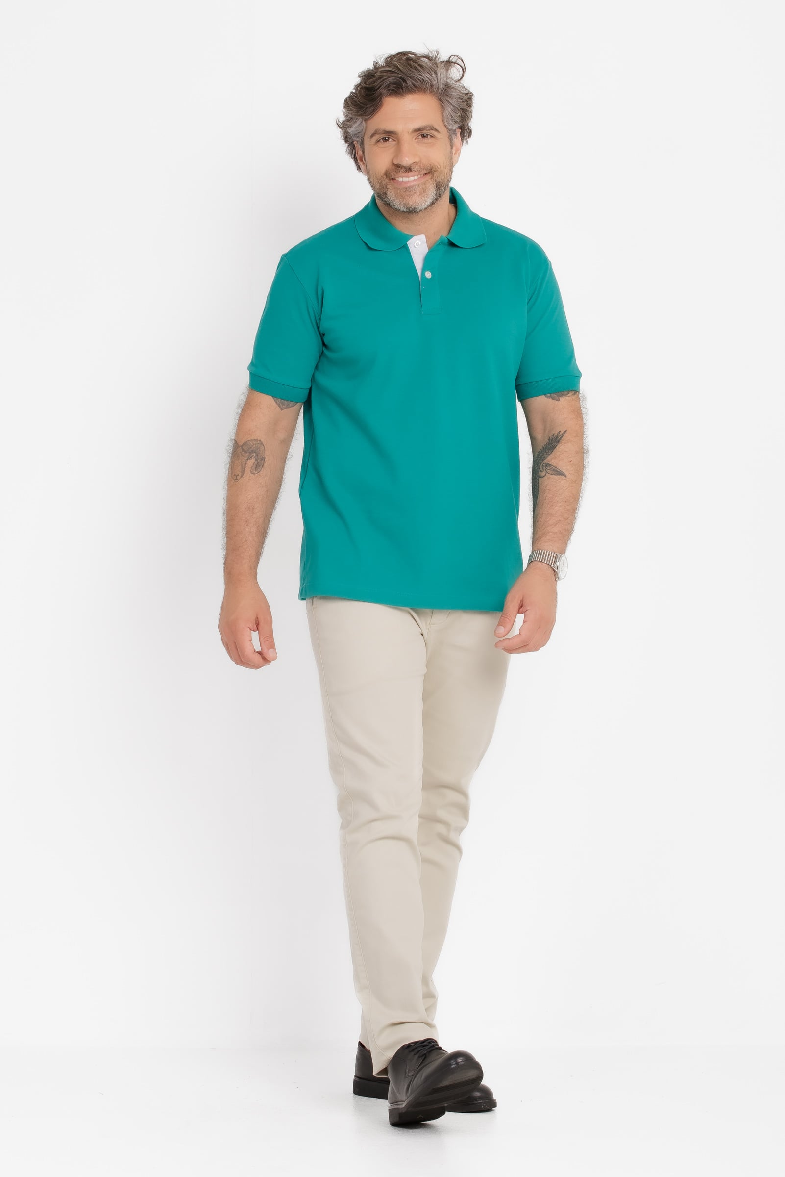 Homem adulto fotografado em estúdio com fundo branco, em enquadramento de corpo inteiro caminhando em direção à câmera. Ele veste camisa polo verde água de manga curta, com gola tradicional e carcela com dois botões claros, em malha piquê de textura uniforme e caimento reto. Usa calça em tom bege claro e sapatos sociais pretos. O modelo tem cabelo ondulado com fios grisalhos, barba curta grisalha e expressão sorridente e confiante. Tatuagens aparecem nos antebraços e há um relógio metálico no pulso esquerdo. Iluminação homogênea e estética clean típica de fotografia de catálogo de moda masculina.