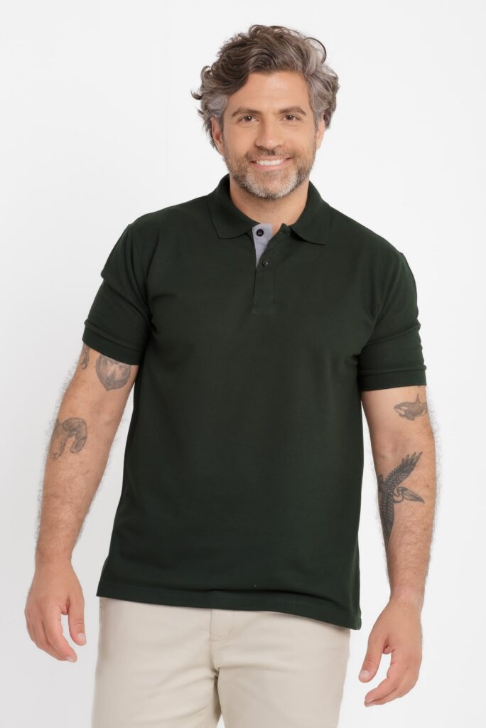 Homem adulto em estúdio com fundo branco, enquadramento do tronco até a cabeça, usando camisa polo verde escura de manga curta, com gola estruturada e vista interna em tom cinza claro. O tecido aparenta malha piquê, com caimento reto e acabamento limpo. Ele sorri levemente, tem cabelos grisalhos ondulados e barba curta. Usa calça clara e tem tatuagens visíveis nos dois braços. Iluminação uniforme e neutra, estilo catálogo.
