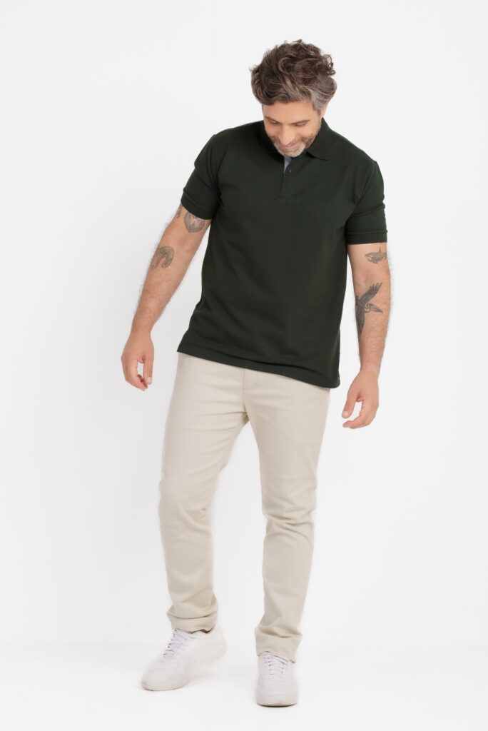 Homem em estúdio com fundo branco usando camisa polo masculina verde escura, de manga curta e caimento ajustado, combinada com calça clara bege e tênis branco, postura relaxada olhando para baixo, com tatuagens visíveis nos braços, visual casual sofisticado que destaca o corte e a simplicidade da peça.