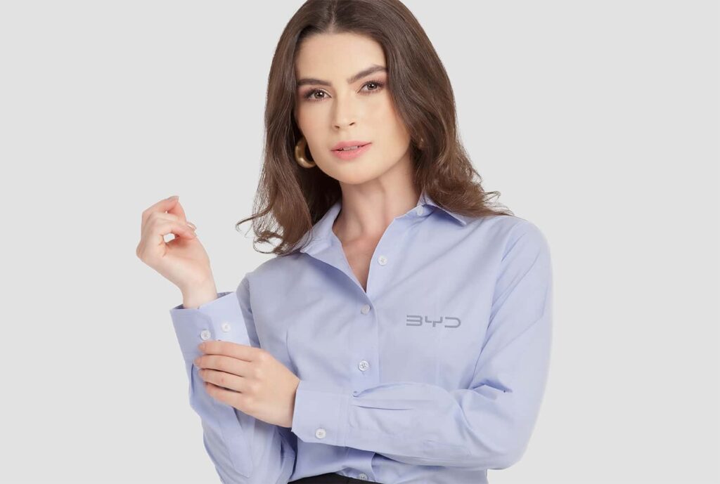 Mulher vestindo camisa social feminina azul clara com logotipo BYD aplicado no lado esquerdo do peito.
