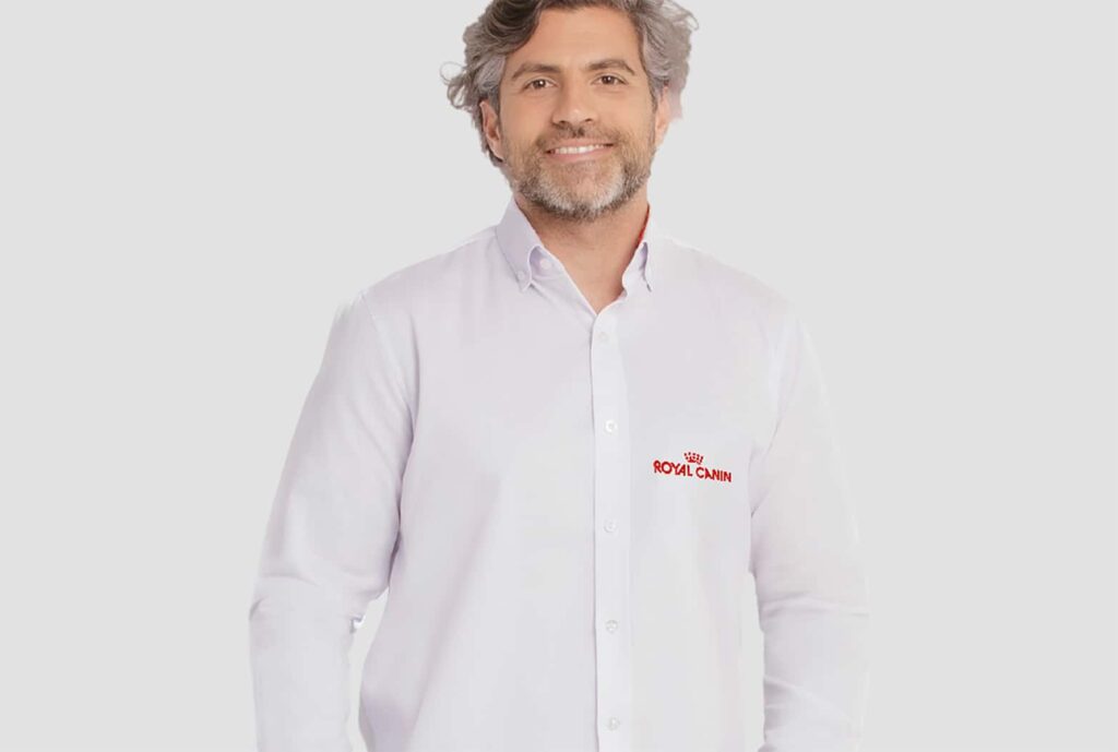 Homem adulto vestindo camisa social branca de manga longa com bordado da marca Royal Canin aplicado no lado esquerdo do peito. A peça possui gola estruturada, fechamento frontal por botões e modelagem alinhada ao corpo. O modelo tem cabelos grisalhos ondulados, barba curta e está posicionado diante de fundo claro neutro.