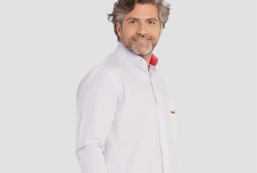 Homem adulto vestindo camisa social branca de manga longa com bordado Royal Canin no lado esquerdo do peito. A peça possui gola estruturada com detalhe interno vermelho contrastante, fechamento frontal por botões e modelagem alinhada. O modelo tem cabelos grisalhos ondulados e barba curta, posicionado em fundo claro neutro.
