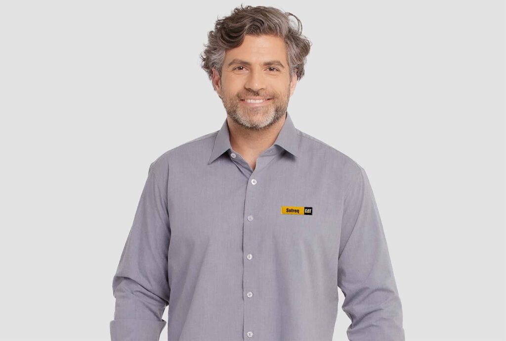 Homem adulto vestindo camisa social cinza de manga longa com bordado Sotreq CAT aplicado no lado esquerdo do peito. A peça possui gola clássica estruturada, fechamento frontal por botões claros e modelagem alinhada ao corpo. O modelo tem cabelos grisalhos ondulados, barba curta e está posicionado diante de fundo claro neutro.