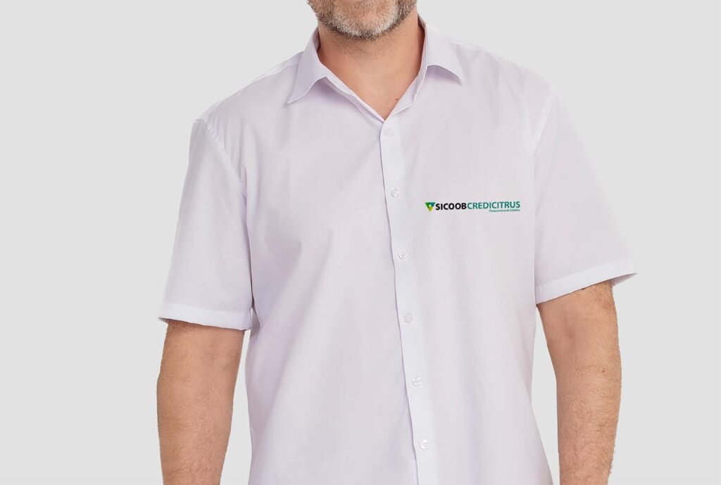 Homem vestindo camisa social branca de manga curta com bordado Sicoob Credicitrus aplicado no lado esquerdo do peito. A peça possui gola clássica, fechamento frontal por botões e modelagem alinhada ao corpo, apresentada em fundo claro neutro.