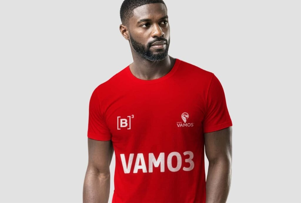 Homem vestindo camiseta vermelha de manga curta com estampa frontal do Grupo Vamos. A peça apresenta a palavra “VAMO3” em destaque na parte inferior, além de logotipos menores aplicados no peito, incluindo símbolo do Grupo Vamos e marca B³.